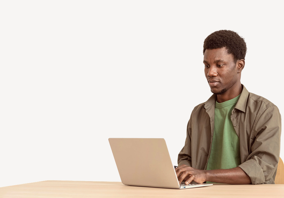 Young man wearing green shirt and brown jacket working on a laptop at a wooden table.